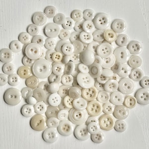 May include: A collection of white and off-white buttons of various sizes and styles. The buttons are arranged in a circular pattern on a white surface.