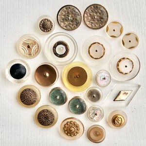 May include: A collection of vintage buttons in various shapes, sizes, and materials. The buttons include clear, gold, brown, and iridescent designs. Some buttons have intricate patterns and textures, while others are smooth and simple.