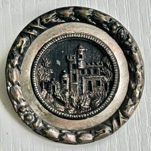 May include: A round, antique button with a detailed castle design. The button features a silver-colored outer rim with a floral pattern, surrounding a dark gray center with the castle illustration. The castle has multiple towers and is surrounded by trees.
