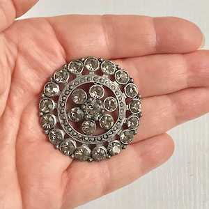 May include: A silver-toned brooch with a circular design featuring a central cluster of clear rhinestones surrounded by a ring of smaller rhinestones.