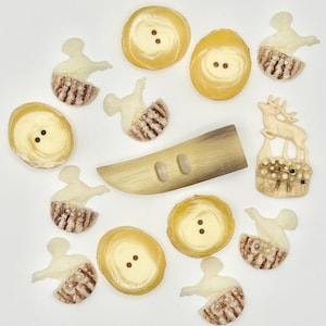 May include: A collection of vintage buttons in various shapes and designs. The assortment includes round buttons with two holes, decorative bird and deer-shaped buttons, and a large, horn-shaped button. The buttons are primarily cream and brown.