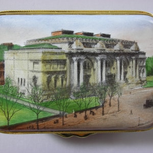 May include: A vintage enamel box with a painted image of a large building with columns and a green roof. The building is surrounded by trees and a street with a car and people.