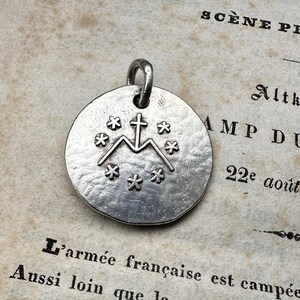May include: A circular silver-toned metal pendant featuring a textured surface. The center displays an embossed design of a cross resting on the letter M, surrounded by a circular arrangement of seven small six-pointed star symbols.