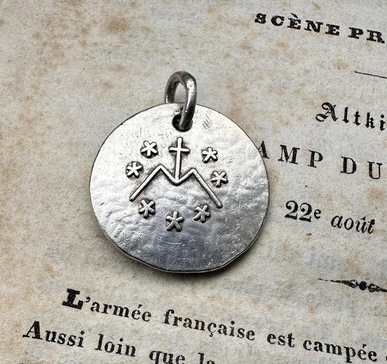 May include: A circular silver-toned metal pendant featuring a textured surface. The center displays an embossed design of a cross resting on the letter M, surrounded by a circular arrangement of seven small six-pointed star symbols.