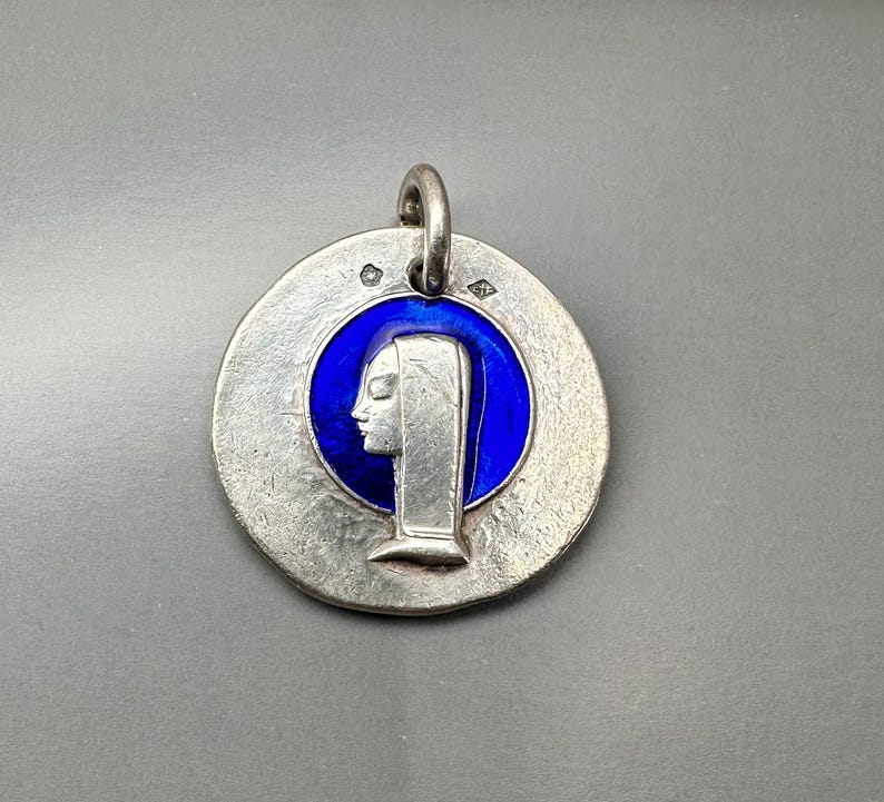 May include: A circular silver pendant featuring a central deep blue enamel inlay. The silver surface has a textured, matte finish and displays a raised profile of a figure wearing a head covering. A small silver bail is attached to the top edge.