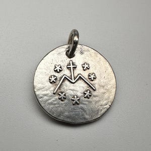 May include: A circular silver-toned metal pendant with a textured, uneven surface. The center features an embossed letter M topped with a cross, surrounded by a circular arrangement of six small, six-pointed star symbols. A small metal loop is attached.