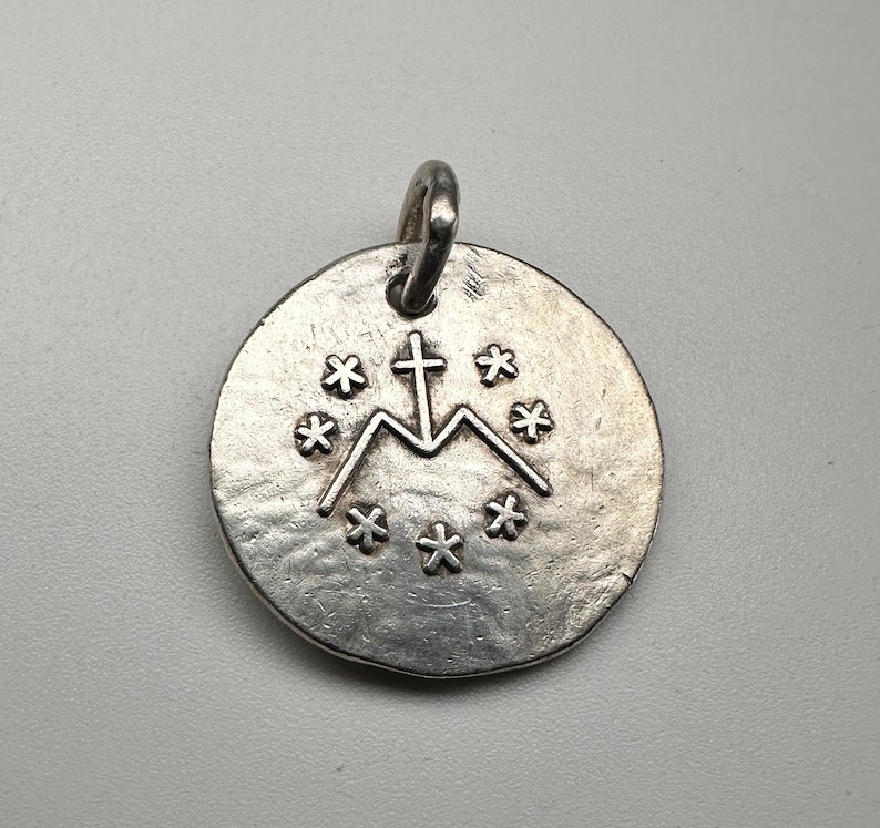 May include: A circular silver-toned metal pendant with a textured, uneven surface. The center features an embossed letter M topped with a cross, surrounded by a circular arrangement of six small, six-pointed star symbols. A small metal loop is attached.