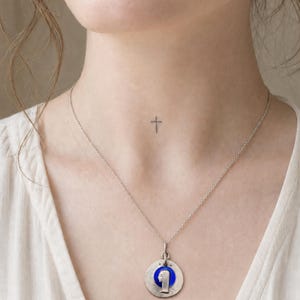 May include: A silver-toned necklace featuring a circular pendant with a textured metallic finish. The center of the pendant contains a vibrant blue circular inlay with a raised silver-toned profile silhouette of a figure facing to the left.
