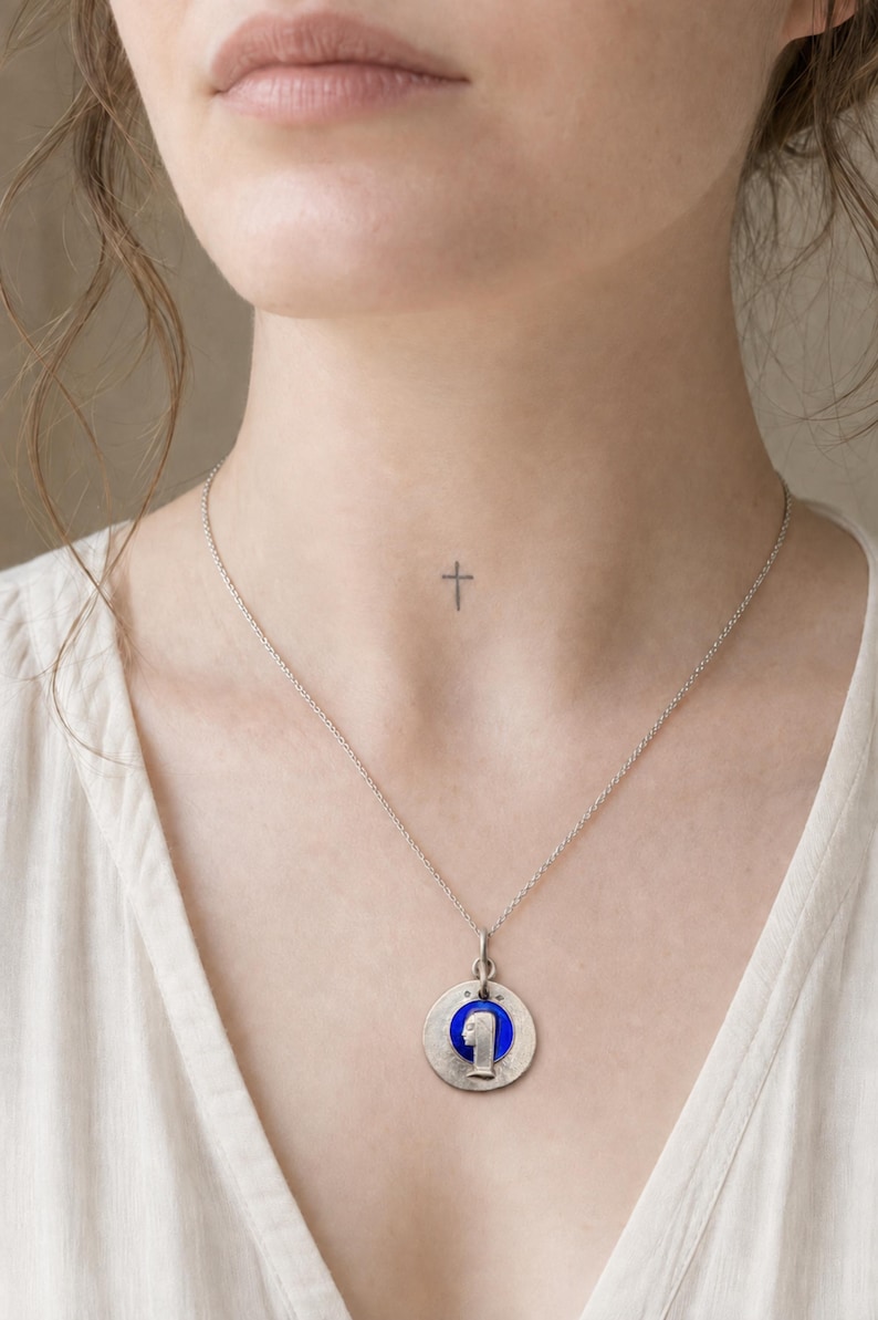 May include: A silver-toned necklace featuring a circular pendant with a textured metallic finish. The center of the pendant contains a vibrant blue circular inlay with a raised silver-toned profile silhouette of a figure facing to the left.