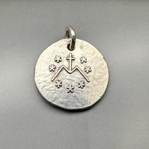 May include: A circular silver-toned metal pendant featuring a textured surface. The center displays an embossed design of a letter M surmounted by a cross, surrounded by a circular arrangement of six small six-pointed star symbols.