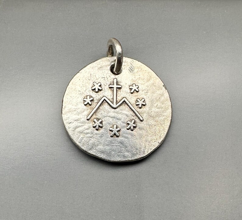 May include: A circular silver-toned metal pendant featuring a textured surface. The center displays an embossed design of a letter M surmounted by a cross, surrounded by a circular arrangement of six small six-pointed star symbols.
