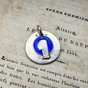 May include: A circular silver pendant featuring a central blue enamel inlay with a profile relief of a figure wearing a head covering. The pendant has a small metal loop at the top for attachment and a textured, matte silver finish on the surface.