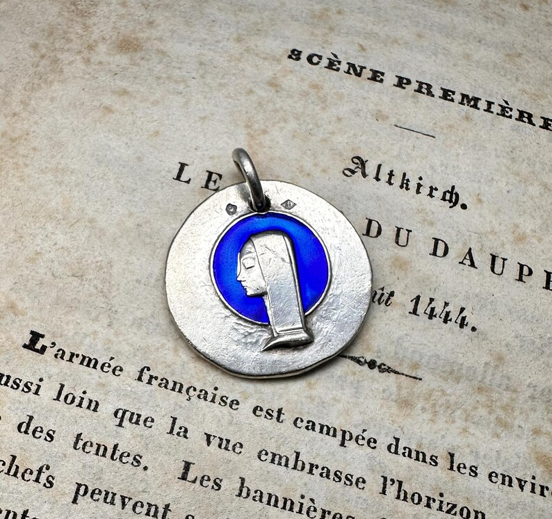 May include: A circular silver pendant featuring a central blue enamel inlay with a profile relief of a figure wearing a head covering. The pendant has a small metal loop at the top for attachment and a textured, matte silver finish on the surface.