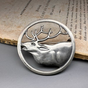 May include: A silver-toned metal pin in the shape of a deer's head with antlers. The deer's head is in profile, facing the right side of the image. The deer's head is surrounded by a circular frame.