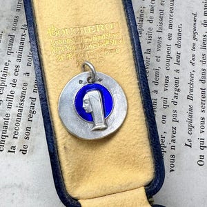 May include: A circular silver pendant featuring a profile relief of a figure wearing a head covering, set against a vibrant blue enamel background. The pendant includes a small silver bail at the top and is presented inside a blue jewelry box.