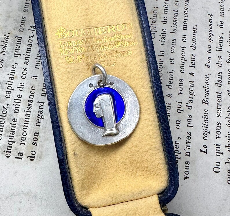 May include: A circular silver pendant featuring a profile relief of a figure wearing a head covering, set against a vibrant blue enamel background. The pendant includes a small silver bail at the top and is presented inside a blue jewelry box.