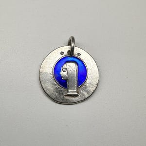 May include: A circular silver pendant featuring a raised profile of a figure wearing a head covering, set against a vibrant blue enamel background. The metal surface has a textured, matte finish with two small hallmarks near the top jump ring.