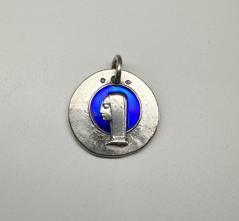 May include: A circular silver pendant featuring a raised profile of a figure wearing a head covering, set against a vibrant blue enamel background. The metal surface has a textured, matte finish with two small hallmarks near the top jump ring.
