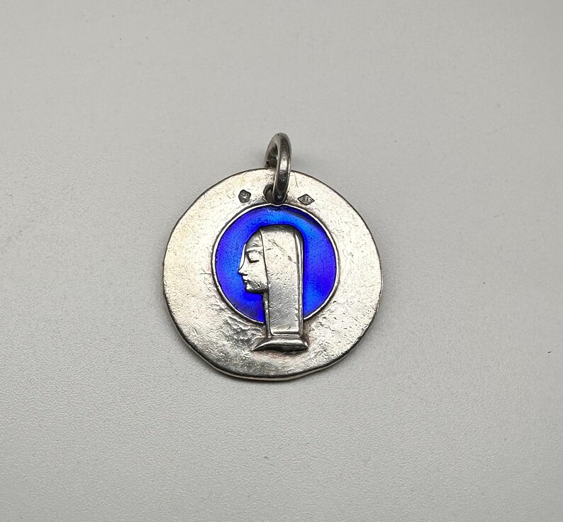 May include: A circular silver pendant featuring a raised profile of a figure wearing a head covering, set against a vibrant blue enamel background. The metal surface has a textured, matte finish with two small hallmarks near the top bail.