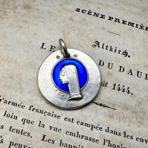 May include: A circular silver-toned metal pendant featuring a raised profile of a person wearing a head covering, set against a vibrant blue enamel circular background. The pendant includes a small metal loop at the top for attachment to a chain.