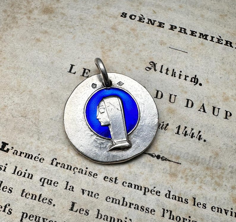 May include: A circular silver-toned metal pendant featuring a raised profile of a person wearing a head covering, set against a vibrant blue enamel circular background. The pendant includes a small metal loop at the top for attachment to a chain.