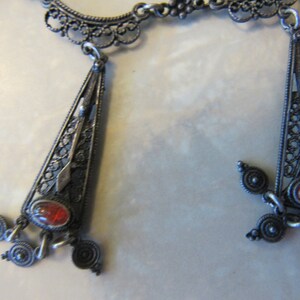 Op de afbeelding: Een zilveren filigraanketting met twee hangende hangers. Elke hanger heeft een rode edelsteen en ingewikkelde filigraanontwerpen.