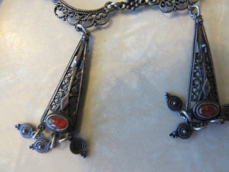 Op de afbeelding: Een zilveren filigraanketting met twee hangende hangers. Elke hanger heeft een rode edelsteen en ingewikkelde filigraanontwerpen.