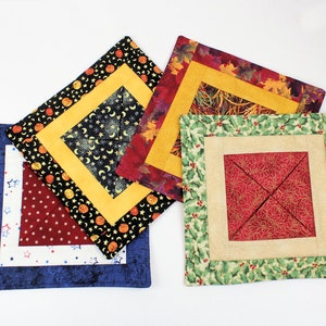 Peut inclure: Quatre dessous de verre carrés en tissu avec divers motifs. L'un a une bordure bleue avec des étoiles blanches, un autre une bordure noire avec des citrouilles et des lunes, un troisième un motif de feuilles d'automne et le dernier un motif de houx et de baies.