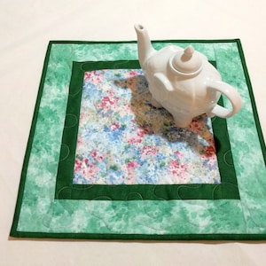 May include: A white ceramic teapot sits on a square quilted placemat. The placemat features a floral pattern in the center, surrounded by a dark green border and a light green outer edge. The teapot has a curved spout and a small lid.