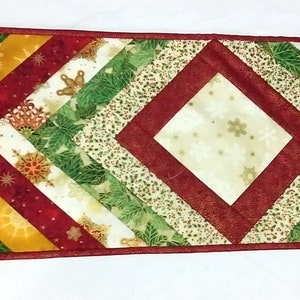 Op de afbeelding: Een rechthoekige, doorgestikte tafelloper met een geometrisch ruitpatroon. De stof heeft een feestelijk kleurenpalet van rood, donkergroen, goud en crème, met bedrukte motieven van sneeuwvlokken, hulstbladeren en zwierige botanische patronen.