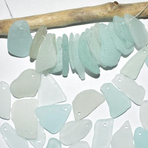 May include: A collection of sea glass pieces in various shapes and sizes, ranging from clear to light blue. The sea glass pieces have holes drilled in them, suggesting they are intended for crafting.