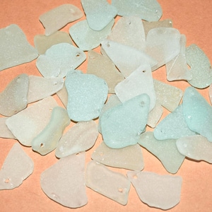 May include: A selection of sea glass pieces in assorted shapes and sizes, in pale green, white, and beige tones. Each piece has a small hole, suitable for jewellery or craft making. The pieces are scattered on a peach-coloured surface.