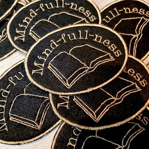 May include: Close-up of several black felt patches with gold embroidery. Each patch is oval-shaped and features an open book with the text "Mind-full-ness" in a gold script font.