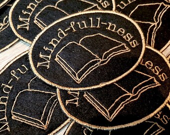 Peut inclure: Gros plan sur plusieurs patchs en feutre noir avec des broderies dorées. Chaque patch est de forme ovale et présente un livre ouvert avec le texte "Mind-full-ness" en écriture dorée.