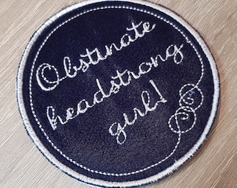 May include: A navy blue round patch with white embroidery that reads "Obstinate headstrong girl!"