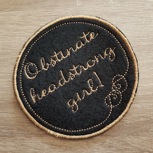 May include: A black round patch with gold stitching. The text "Obstinate headstrong girl" is embroidered in gold on the patch.