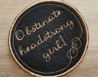 May include: A black round patch with gold stitching. The text "Obstinate headstrong girl" is embroidered in gold on the patch.