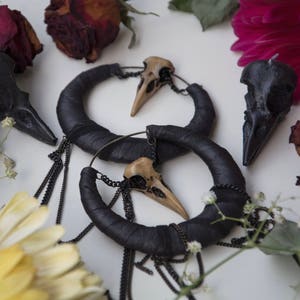 May include: A pair of black hoop earrings with a skull charm hanging from each hoop. The earrings are surrounded by dried flowers and a yellow daisy.