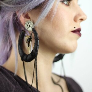 May include: A person with lavender hair wearing large black leather hoop earrings with silver and bone accents and a chain dangle.