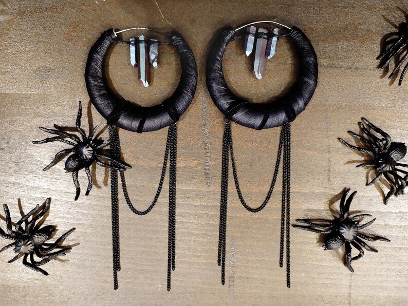 May include: A pair of large black hoop earrings with dangling chains and crystal accents. The earrings are on a wooden surface with black plastic spiders.