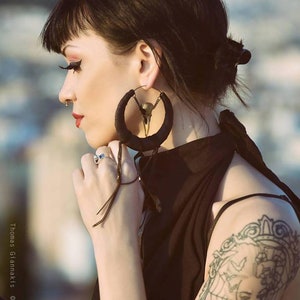 May include: A woman with black hair wearing large black hoop earrings with a skull design. The earrings have a long tassel hanging from them. She is wearing a black sleeveless top and has a tattoo on her upper arm.