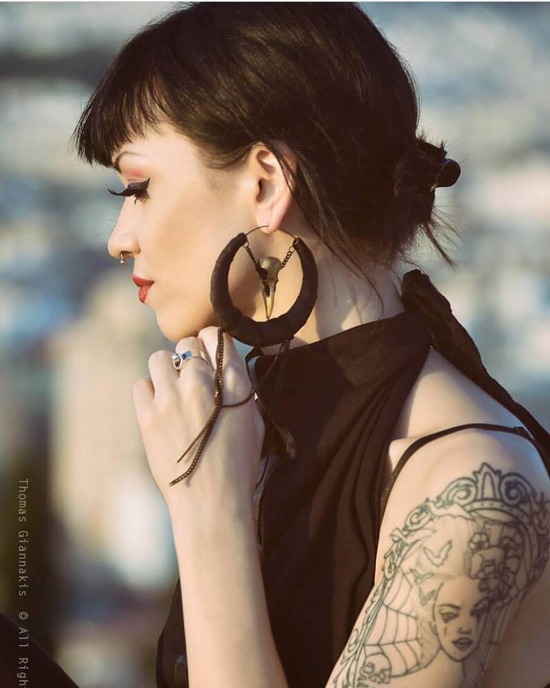 May include: A woman with black hair wearing large black hoop earrings with a skull design. The earrings have a long tassel hanging from them. She is wearing a black sleeveless top and has a tattoo on her upper arm.