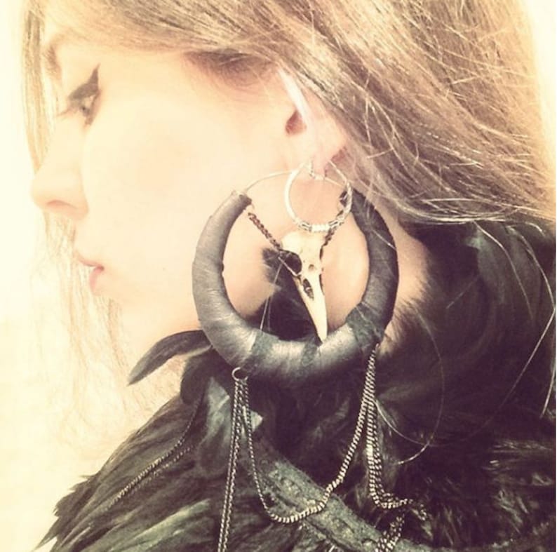 May include: Large black hoop earrings with a bird skull charm and feather accents. The earrings are made of leather and have a silver hoop.