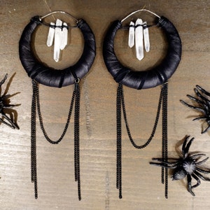 May include: A pair of large hoop earrings with black fabric wrapped around the hoops. Each hoop has a cluster of clear quartz crystals hanging from it. The earrings have multiple black chains hanging from the bottom.