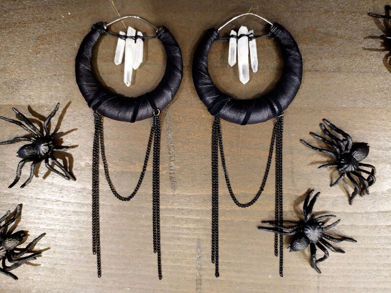 May include: A pair of large hoop earrings with black fabric wrapped around the hoops. Each hoop has a cluster of clear quartz crystals hanging from it. The earrings have multiple black chains hanging from the bottom.