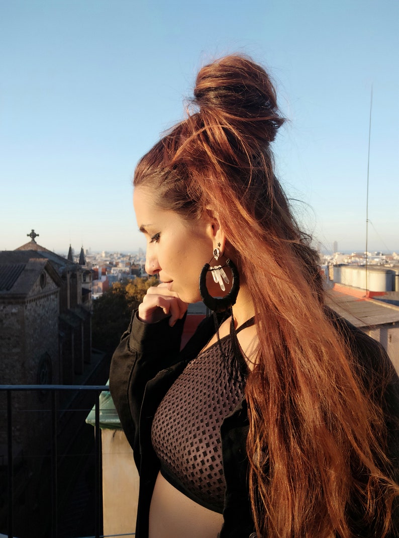 May include: A woman with long, red hair styled in a messy bun wears a black crop top with a mesh design and a black jacket. She is wearing large hoop earrings with white crystal accents.