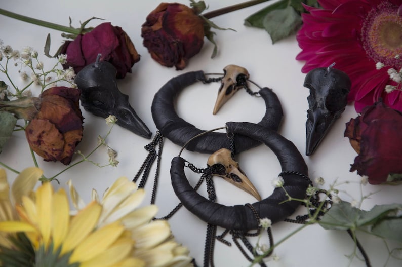 May include: A pair of black hoop earrings with a skull charm hanging from each hoop. The earrings are surrounded by dried flowers and a yellow daisy.