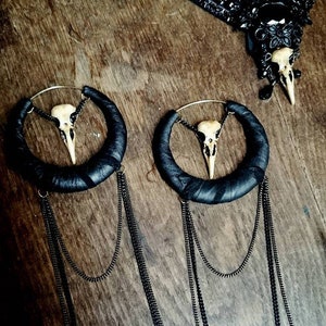 May include: A pair of black leather hoop earrings with a small bird skull charm hanging from each hoop. The earrings have multiple black chain tassels hanging from the bottom.