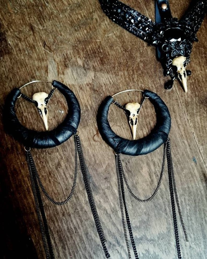 May include: A pair of black leather hoop earrings with a small bird skull charm hanging from each hoop. The earrings have multiple black chain tassels hanging from the bottom.