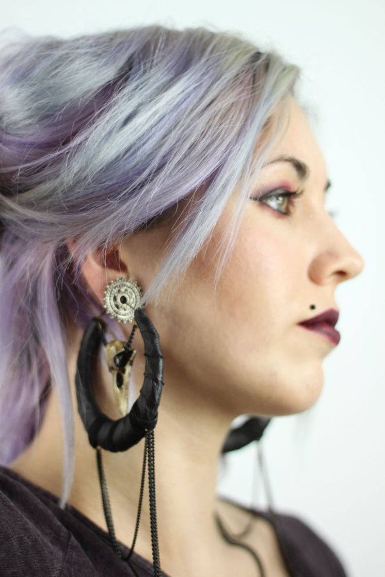 May include: A person with lavender hair wearing large black leather hoop earrings with silver and bone accents and a chain dangle.